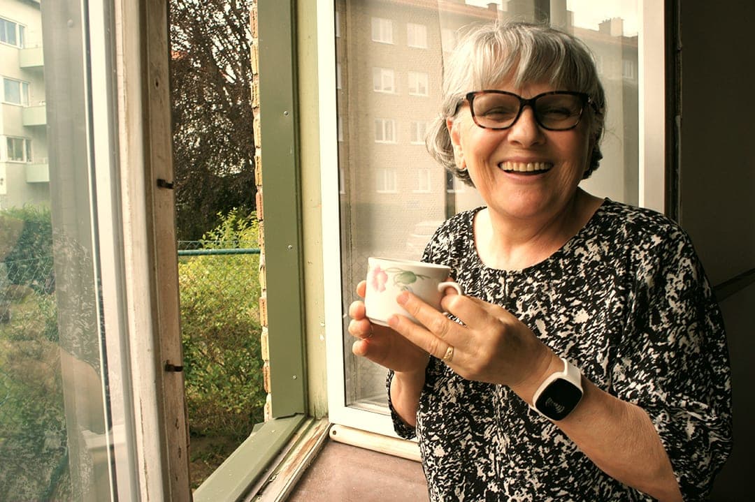 Eine lächelnde Frau, die TM-Alert an ihrem Handgelenk trägt und eine Tasse an einem offenen Fenster hält