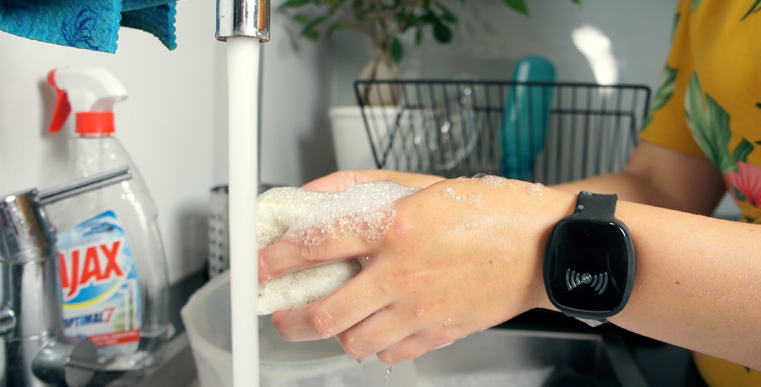 Close-up of a person washing dishes at a kitchen sink with water running and soap suds on their hands, while wearing a black TM-Alert smart wristband.