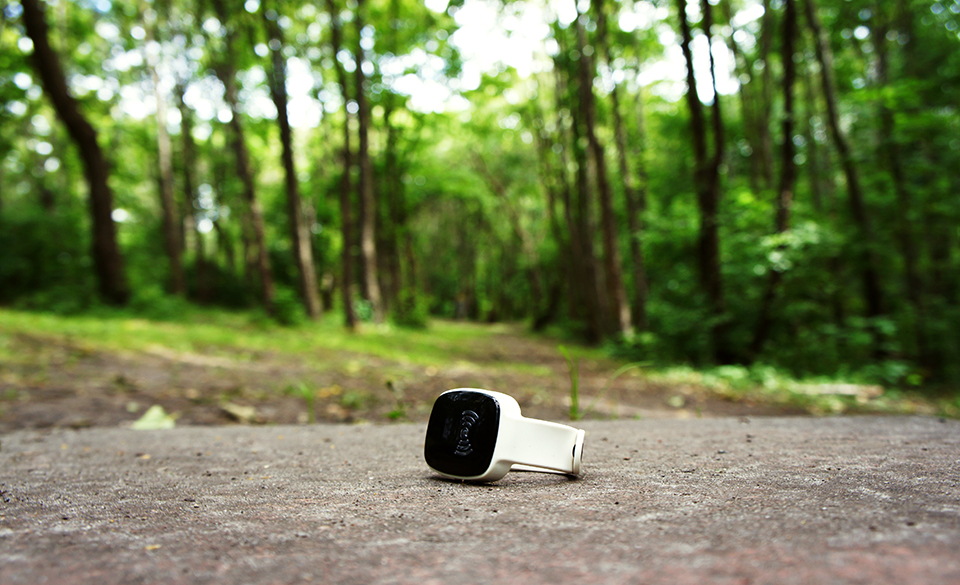 A close-up of a wearable alert device placed on a concrete surface in a lush forest setting. The band is white and the face of the device shows a signal icon, contrasting sharply with the green, leafy environment in the background.